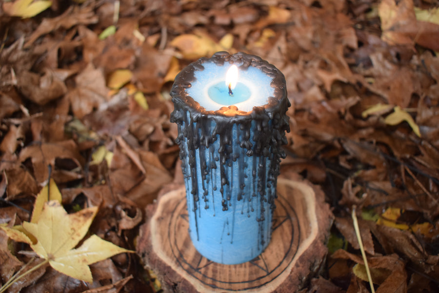 A blue pillar candle with black drips with a flame licking from its wick sits on a pentagram disk resting on a bed of leaves