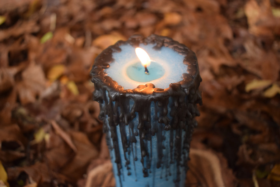 A blue pillar candle with black drips with a flame licking from its wick resting on a bed of leaves