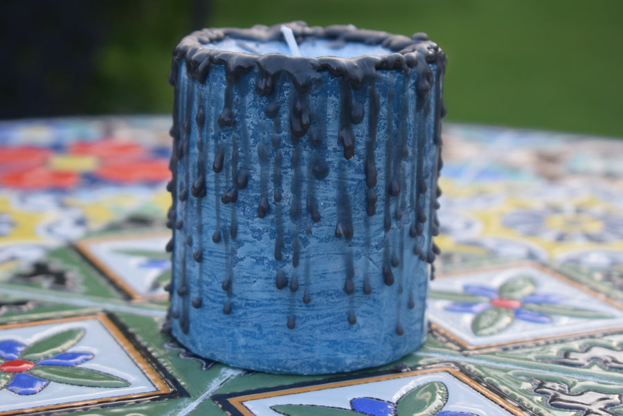 A blue pillar candle with black drips sits on a mosaic table.