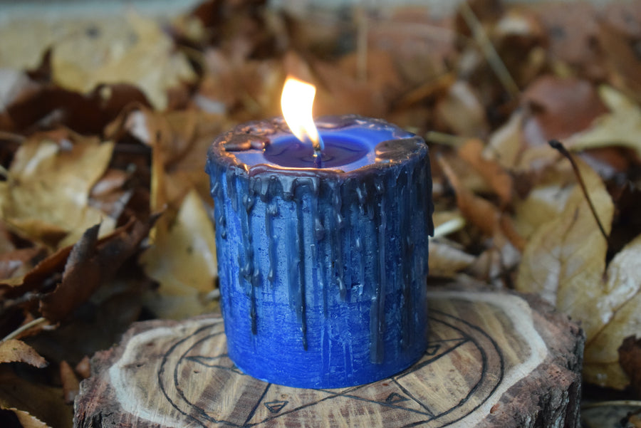 A blue pillar candle with black drips sits with its wick aflame on a timber pentagram disk nestled on a bed of leaves.