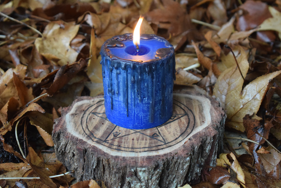 A blue pillar candle with black drips sits aflame on a timber pentagram disk nestled on a bed of leaves.