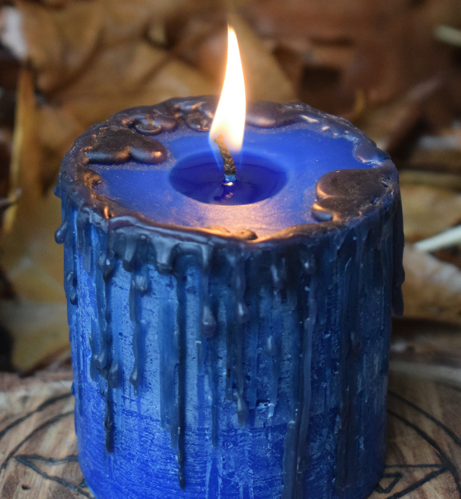 A blue pillar candle with black drips sits with its wick alight on a timber pentagram disk nestled on a bed of leaves.
