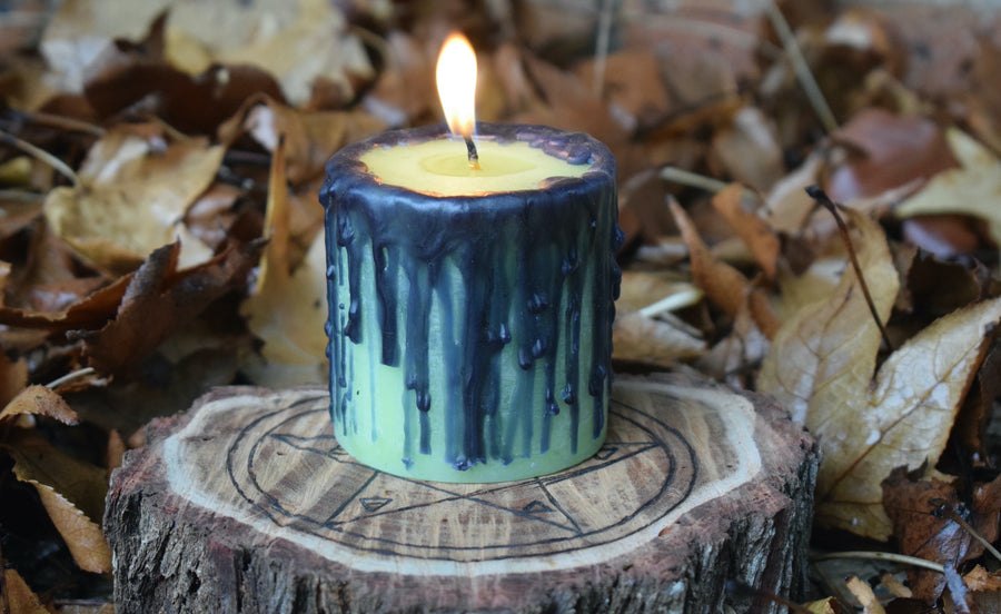 A green pillar candle with black drips sits alight on a pentagram wooden disk nestled on a bed of leaves.