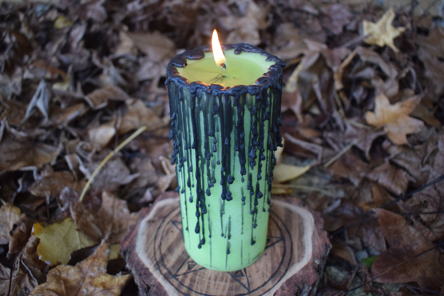 A green pillar candle with black drips sits with its wick alight on a wooden pentagram disk nestled on a bed of autumn leaves.