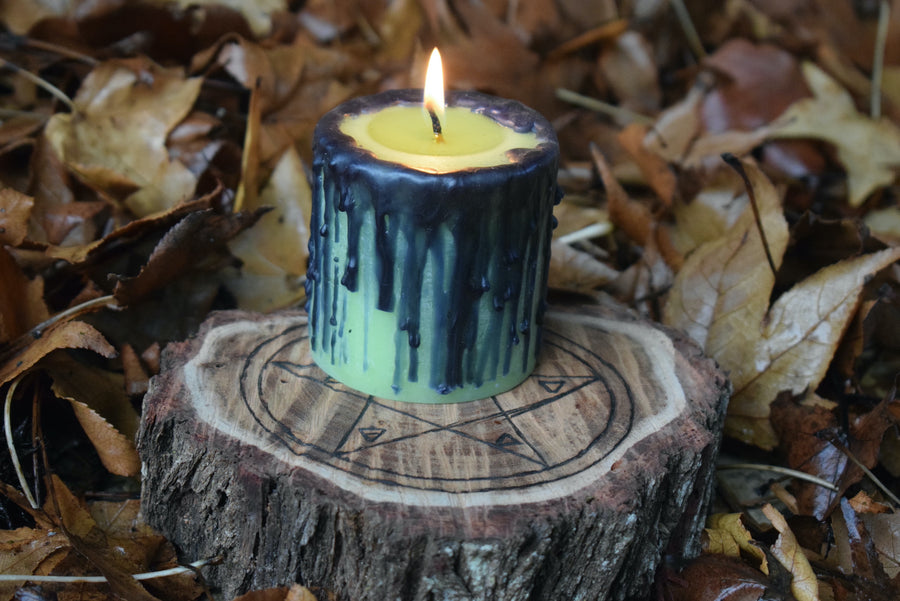 A green pillar candle with black drips sits with its wick lit on a pentagram wooden disk nestled on a bed of leaves.