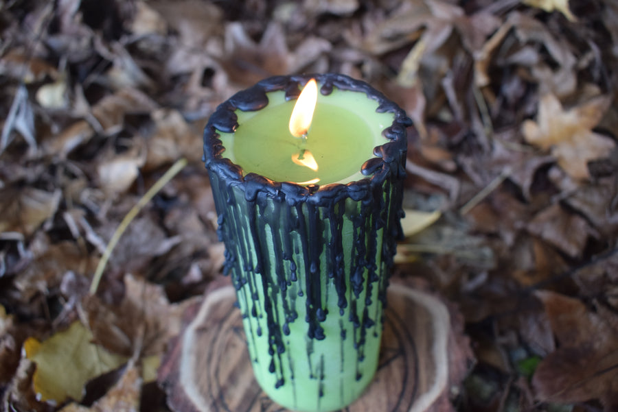 A green pillar candle with black drips sits with its wick lit on a wooden pentagram disk nestled on a bed of autumn leaves.