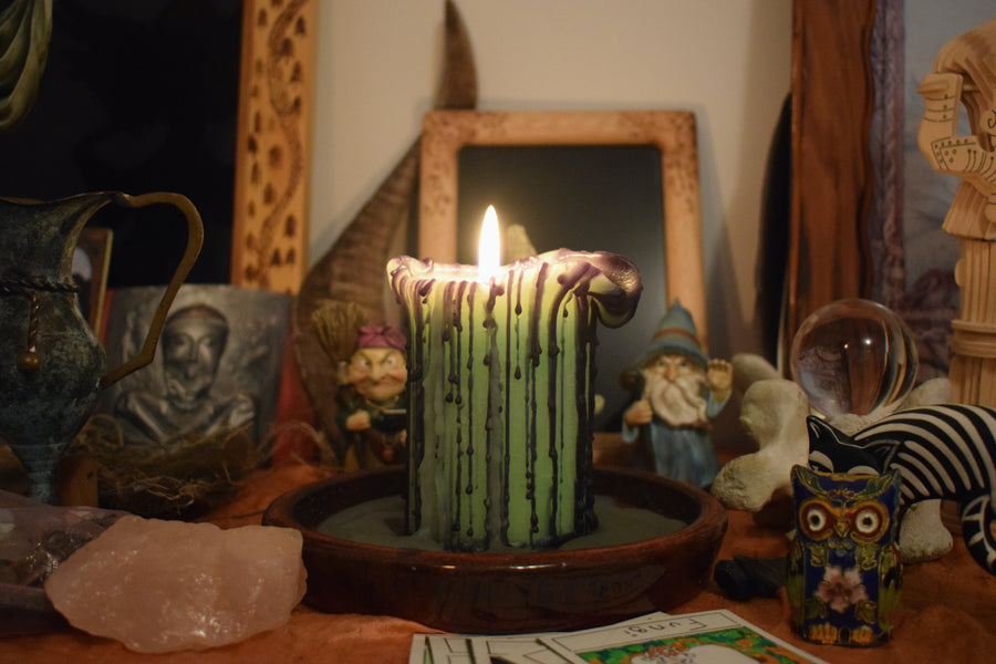 A green pillar candle with black drips sits with its wick aflame on a altar with crystals, ornaments and a crystal ball in the background.