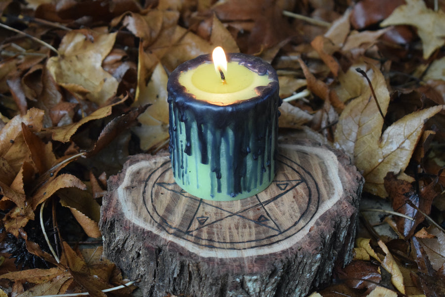 A green pillar candle with black drips sits aflame on a pentagram wooden disk nestled on a bed of leaves.