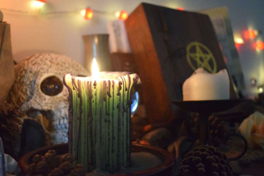 A green pillar candle with black drips sits with its wick alight on a altar with pinecones, a skull and a pentagram grimoire in the background.