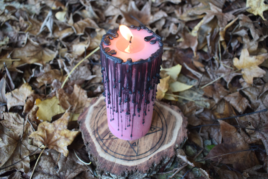 A flame dances from a pink pillar candle with black drips as it rests on a wooden pentagram disk nestled on a bed of autumn leaves