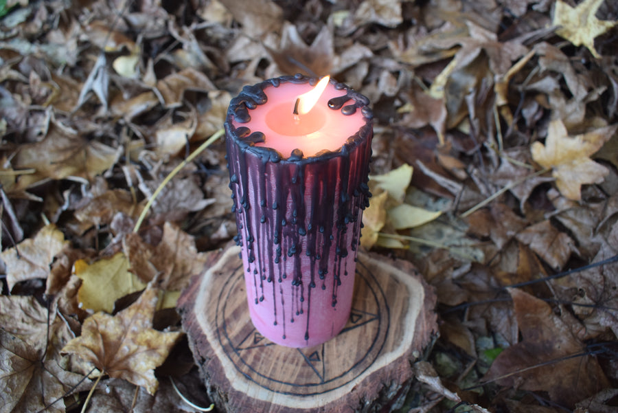 A flame flickers in the breeze from a pink pillar candle with black drips as it rests on a wooden pentagram disk nestled on a bed of autumn leaves
