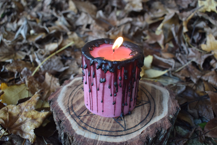 A lit pink pillar candle with a flame dancing from its wick sits on a pentagram disk nestled on a bed of autumn leaves