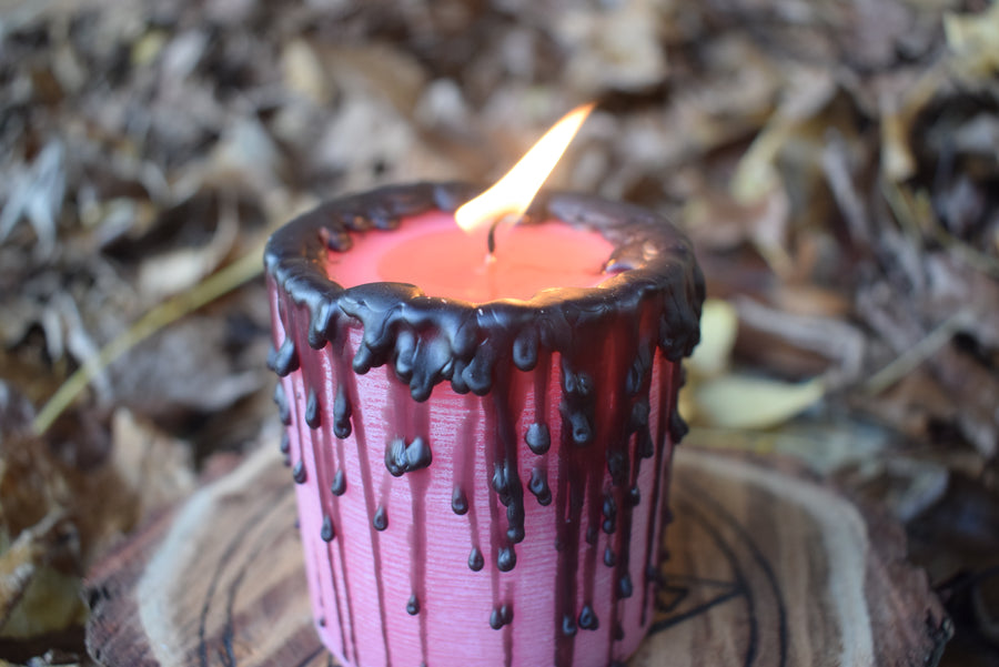 A pink pillar candle with its wick aflame sits on a pentagram disk nestled on a bed of autumn leaves