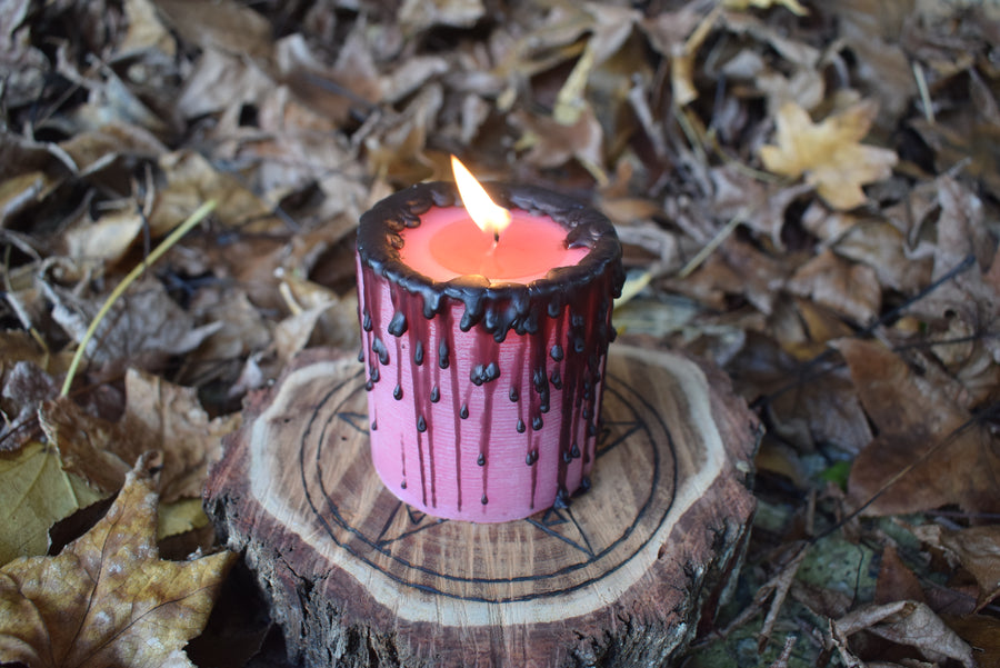 A pink pillar candle with its wick alight sits on a pentagram disk nestled on a bed of autumn leaves