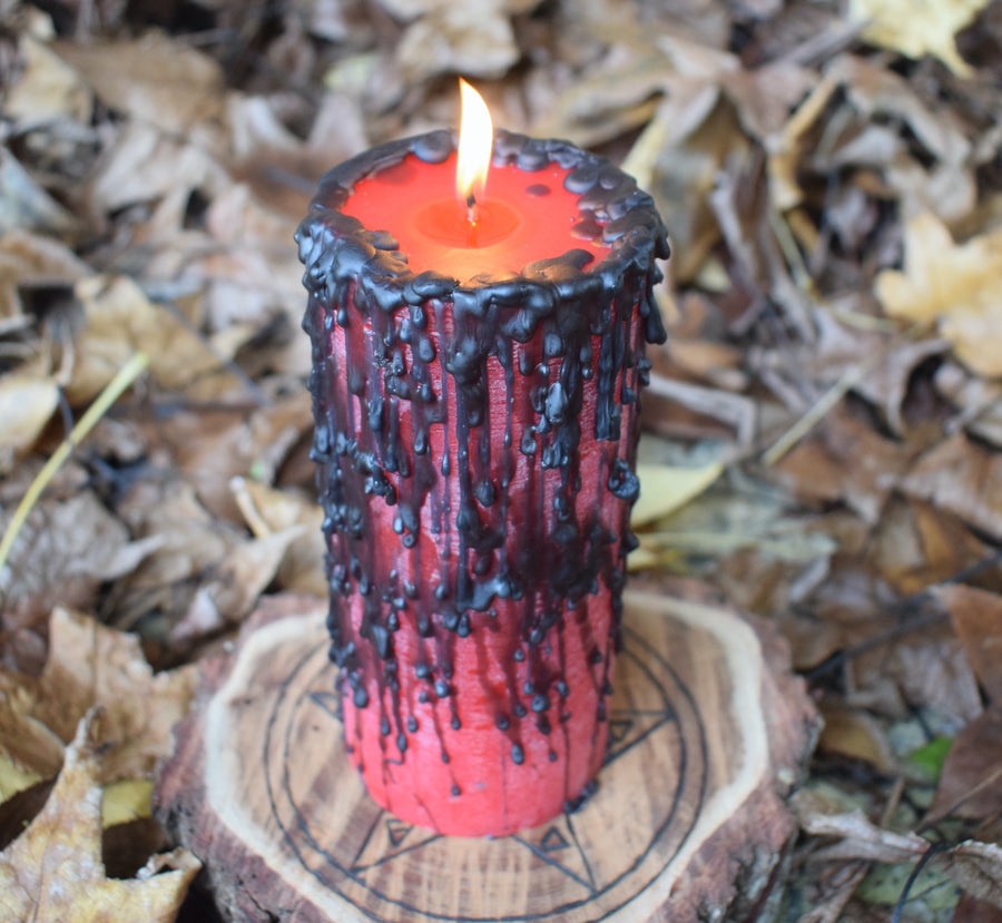 A lit red pillar candle with black wax drips with a flame licking from its wick rests on a pentagram disk nestled on a bed of autumn leaves