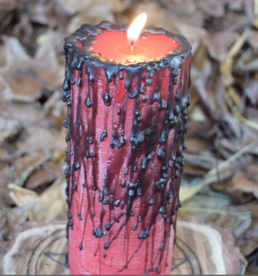 A red pillar candle with black wax drips with a flame licking from its wick rests on a pentagram disk nestled on a bed of autumn leaves