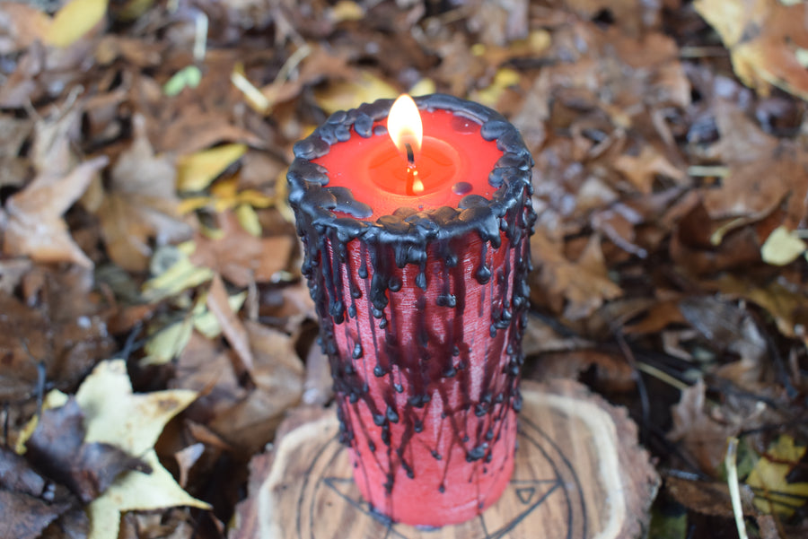 A lit red pillar candle with black wax drips with a flame alight from its wick rests on a pentagram disk nestled on a bed of autumn leaves