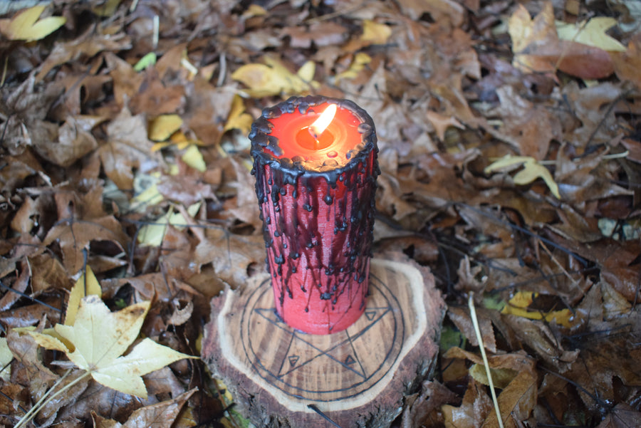 A red pillar candle with black wax drips with a flame licking from its wick rests on a pentagram disk nestled on a bed of autumn leaves