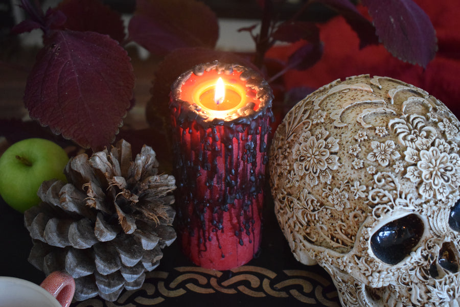A lit red pillar candle with black wax drips with a flame licking from its wick rests on an altar with a skull, pinecone and green apple