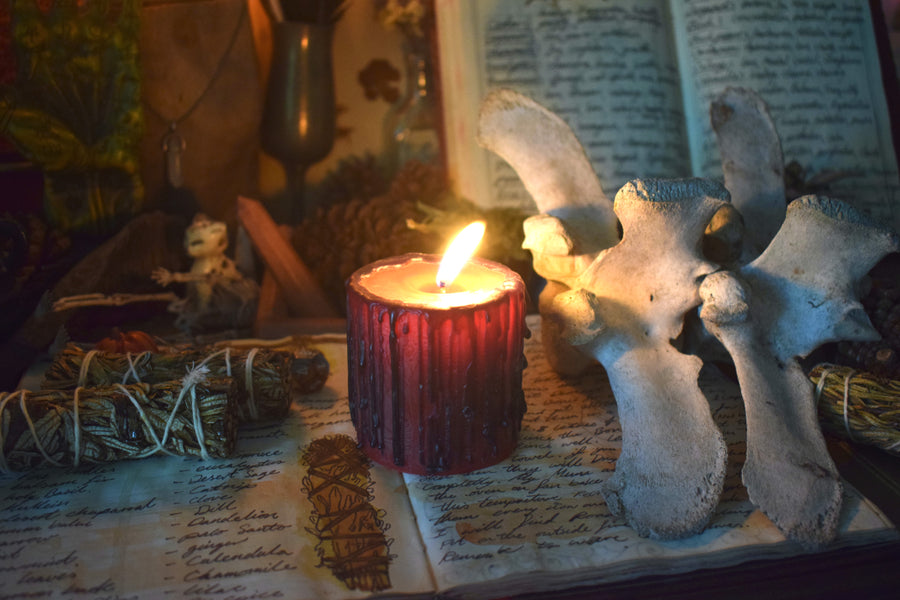 A red candle with black drips sits alight on an open grimoire with bones, smudge sticks and other altarpieces.