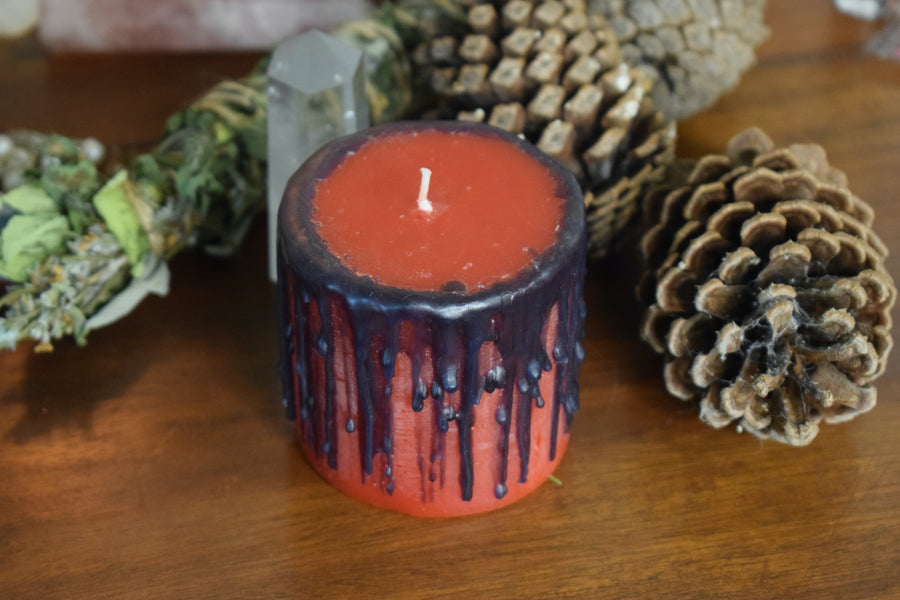 A red candle with black drips sits with pinecones, a crystal and herbs.