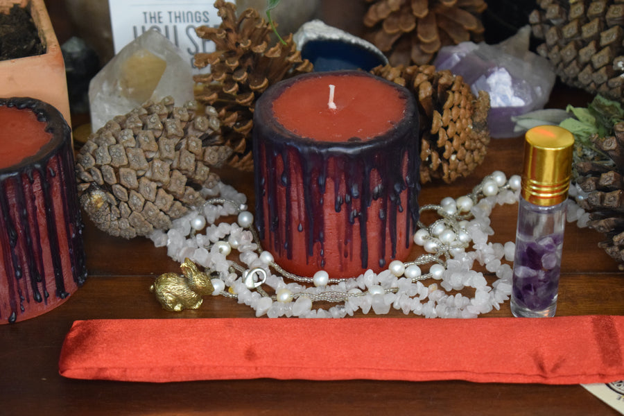 A red candle with black drips sits on an altar with pinecones, beads, a bronze rabbit and pinecones.