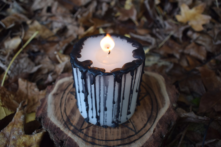 A black and white pillar candle with a flame licking from its wick rests on a wooden pentagram disk on a bed of autumn leaves
