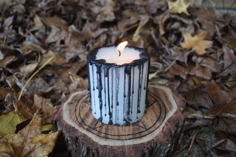 A black and white pillar candle with a flame licking from its wick rests on a wooden pentagram disk on a bed of autumn leaves