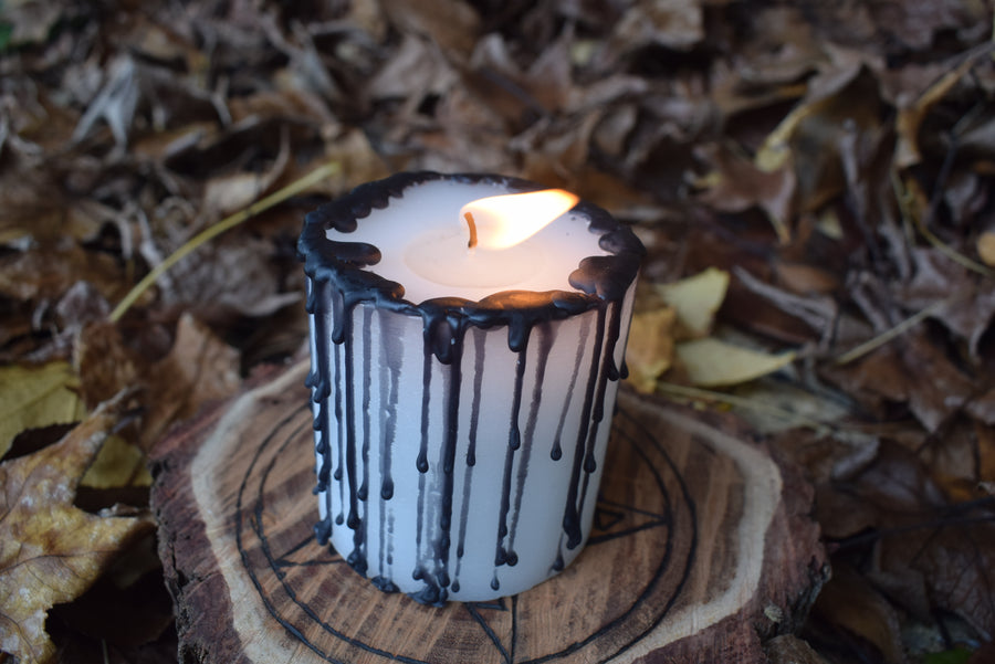 A black and white pillar candle with a flame dances from its wick rests on a wooden pentagram disk on a bed of autumn leaves