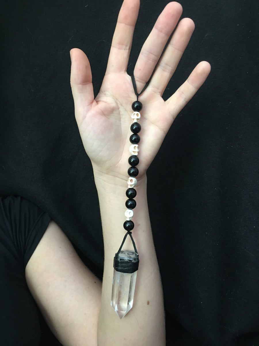 A clear quartz crystal pendulum with skull bone beads and black glass beads on leather resting on an open hand