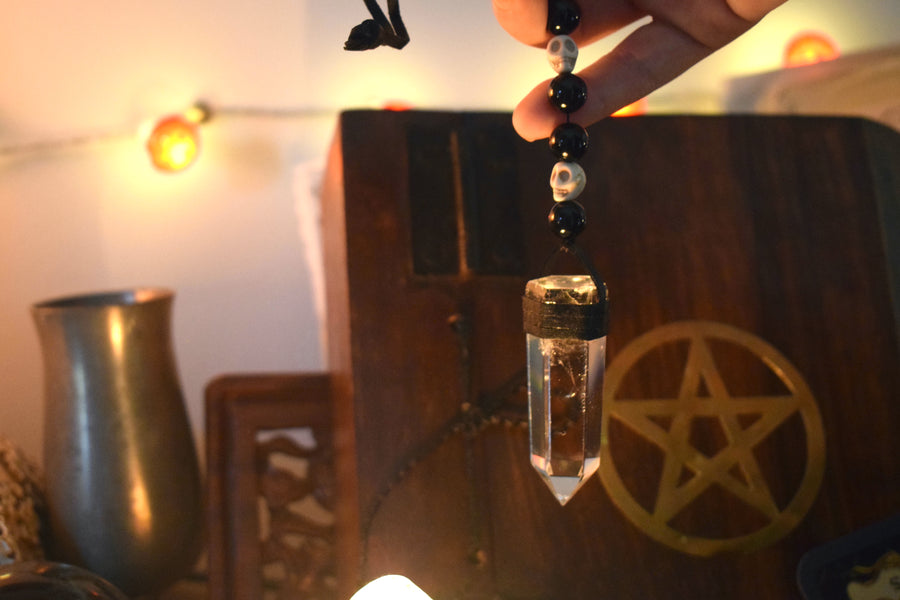 A hand holding a clear quartz crystal pendulum with skull bone beads and black glass beads on leather with a pentagram in the background