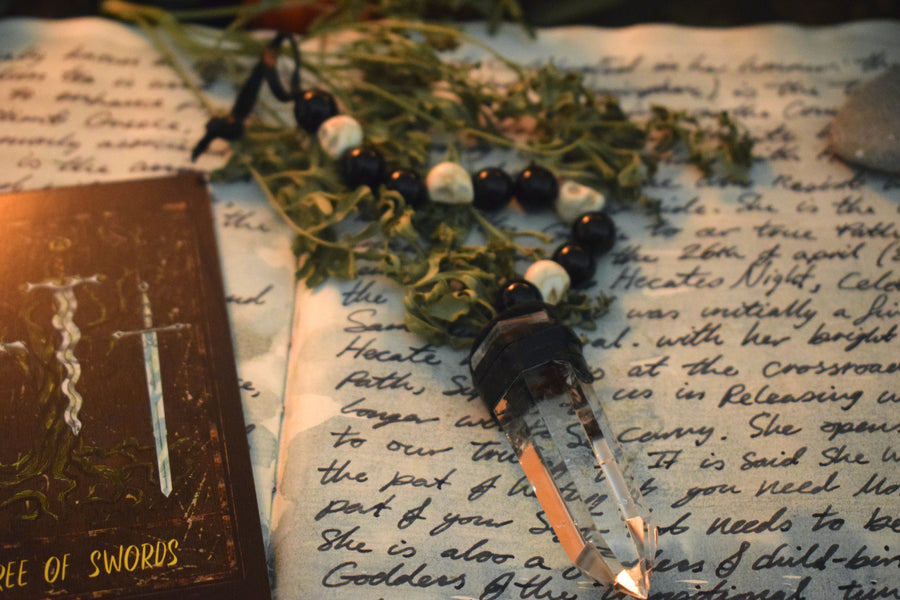 A clear quartz crystal pendulum with skull bone beads and black glass beads on leather resting on an open grimoire with herbs and a tarot card