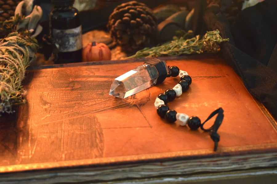 A clear quartz crystal pendulum with skull bone beads and black glass beads on leather resting on a grimoire with herbs and a pinecone in the background