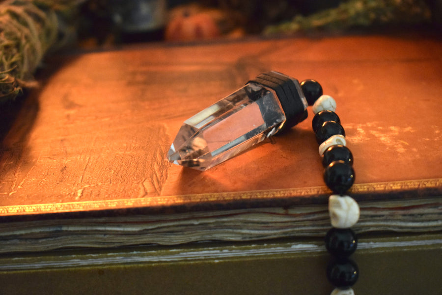 A clear quartz crystal pendulum with skull bone beads and black glass beads on leather resting on a grimoire