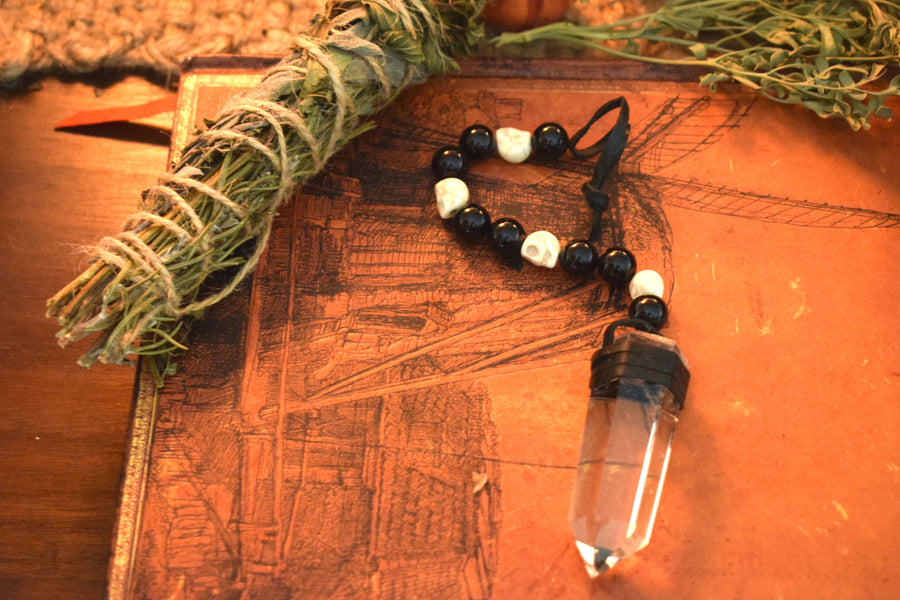 A clear quartz crystal pendulum with skull bone beads and black glass beads on leather resting on a grimoire with herbs