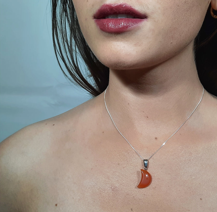 A carnelian crystal crescent moon necklace on a sterling silver chain hanging on the neck of a person