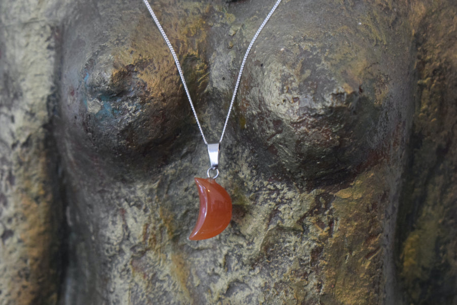 A carnelian crystal crescent moon necklace on a sterling silver chain hanging on the neck of a bronze goddess sculpture
