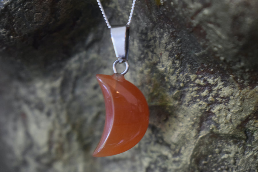A carnelian crystal crescent moon necklace on a sterling silver chain hanging with a bronze sculpture background