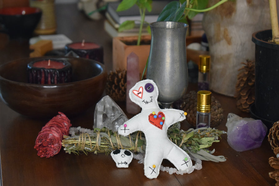 A hoodoo voodoo poppet doll with button eyes and a red love heart on its chest pierced with colour magick pins sits on an altar with pinecones, herbs and a candle.