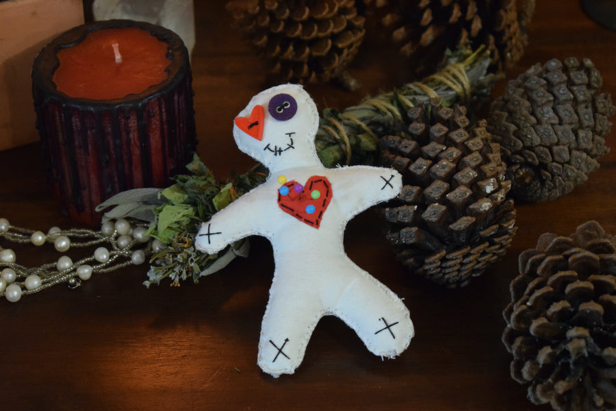 A hoodoo voodoo poppet doll with button eyes and a red love heart on its chest peiced with colour magick pins sits on an altar with pinecones, herbs and a candle.