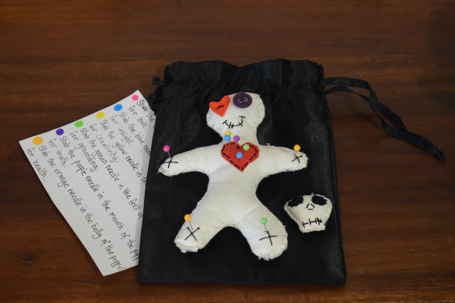 A hoodoo voodoo poppet doll with button eyes and a red love heart on its chest pierced with colour magick pins sits on a black satin drawstring pouch with a skull shaped pin cushion with instructions. 