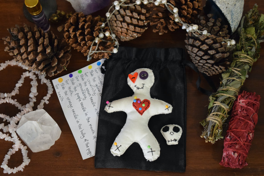 A hoodoo voodoo poppet doll with button eyes and a red love heart on its chest peiced with colour magick pins sits on an altar with a skull pin cushion, a black satin drawstring pouch, instructions, herbs, crystals and pinecones.