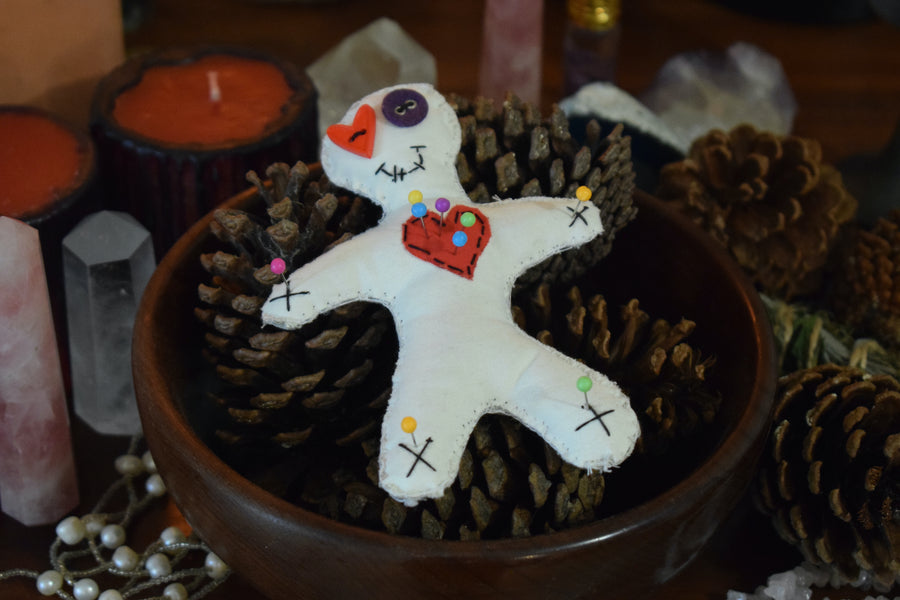 A hoodoo voodoo poppet doll with button eyes and a red love heart on its chest pierced with colour magick pins sits on an altar with pinecones, crystals and a candle.