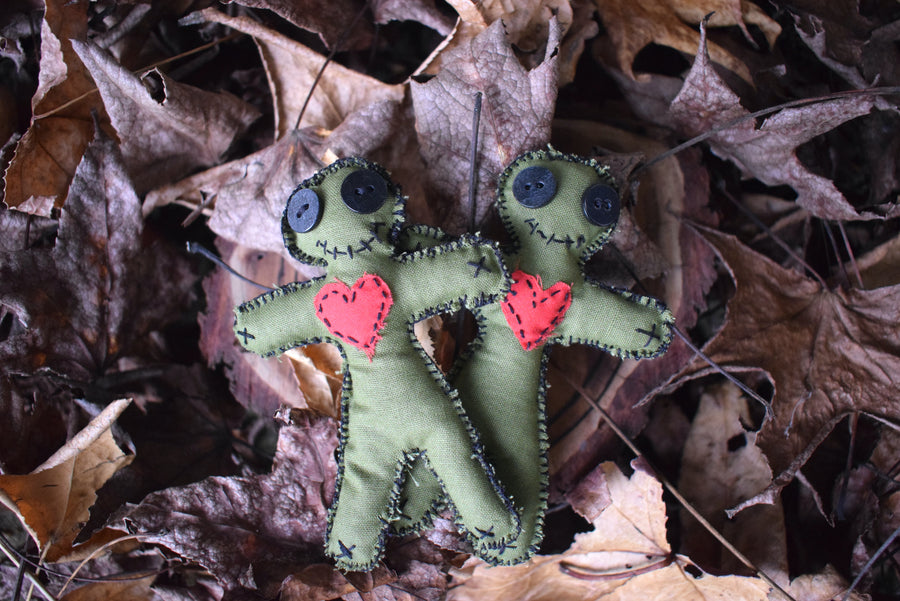 Two green hoodoo voodoo poppet dolls with button eyes, a stitched smile and a red love heart over their chests sit nestled on a bed of autumn leaves.