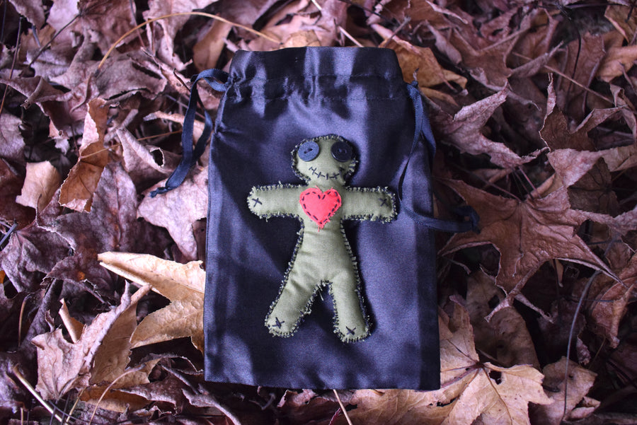 A green hoodoo voodoo poppet doll with button eyes, a stitched smile and a red love heart over its chest sit on a black satin drawstring pouch nestled on a bed of autumn leaves.