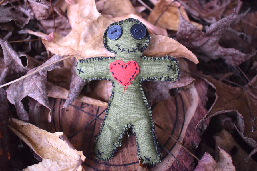 A green hoodoo voodoo poppet doll with button eyes, a stitched smile and a red love heart over their chest sit nestled on a bed of autumn leaves.