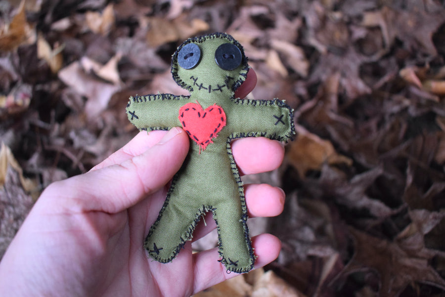 A hand holds a green hoodoo voodoo poppet doll with button eyes, a stitched smile and a red love heart over its chest.