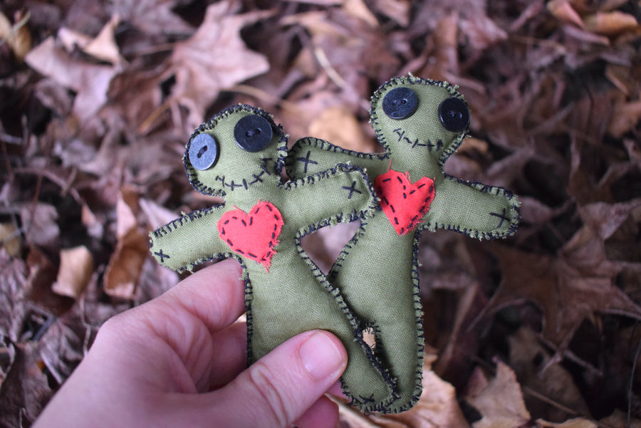 A hand holds two green hoodoo voodoo poppet dolls with button eyes, a stitched smile and a red love heart over their chests with a bed of autumn leaves in the background.