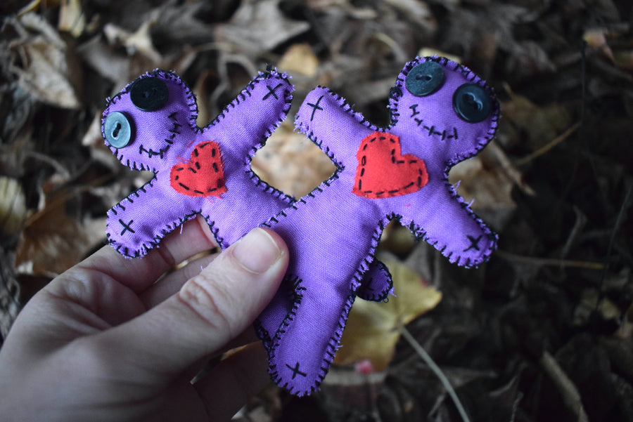 A hand holding two purple fabric hoodoo voodoo dolls with black button eyes, a stitched smile and red fabric love heart on their heart nestled on a bed of leaves.