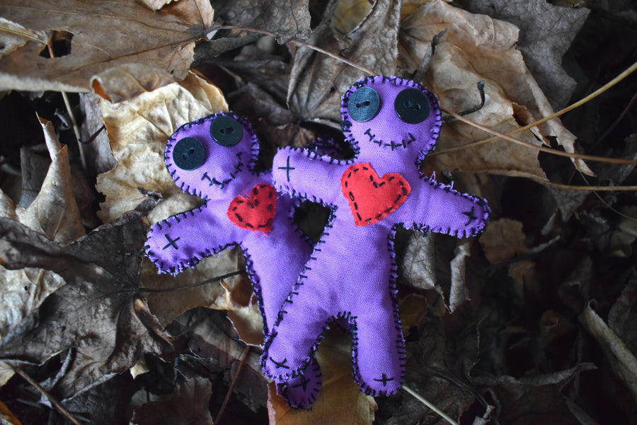 Two purple hoodoo voodoo dolls with black button eyes, a stitched smile and red fabric love heart on their heart nestled on a bed of leaves.